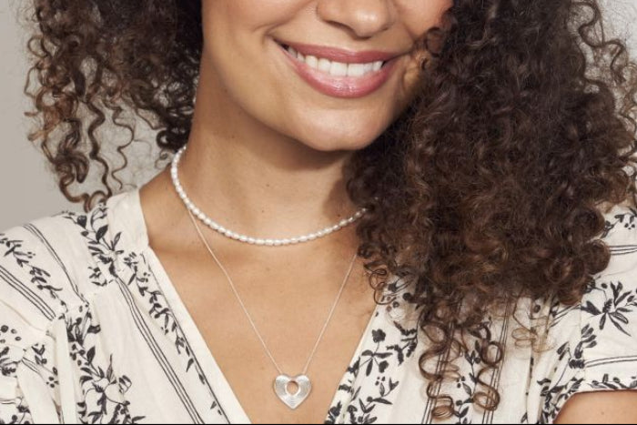 Woman wearing layered necklace style with rice seed pearl choker necklace and silver heart pendant necklace against a neutral background