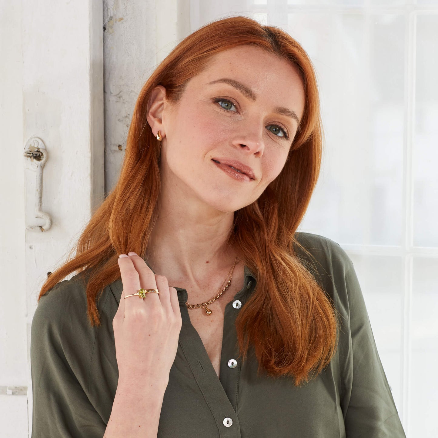 Woman with red hair wearing a green shirt and gold jewellery, on her hand wearing Trilogy Peridot 9ct Gold ring stacked with other gold rings and wearing gold jewellery from Silverage London showing from far. against a white background