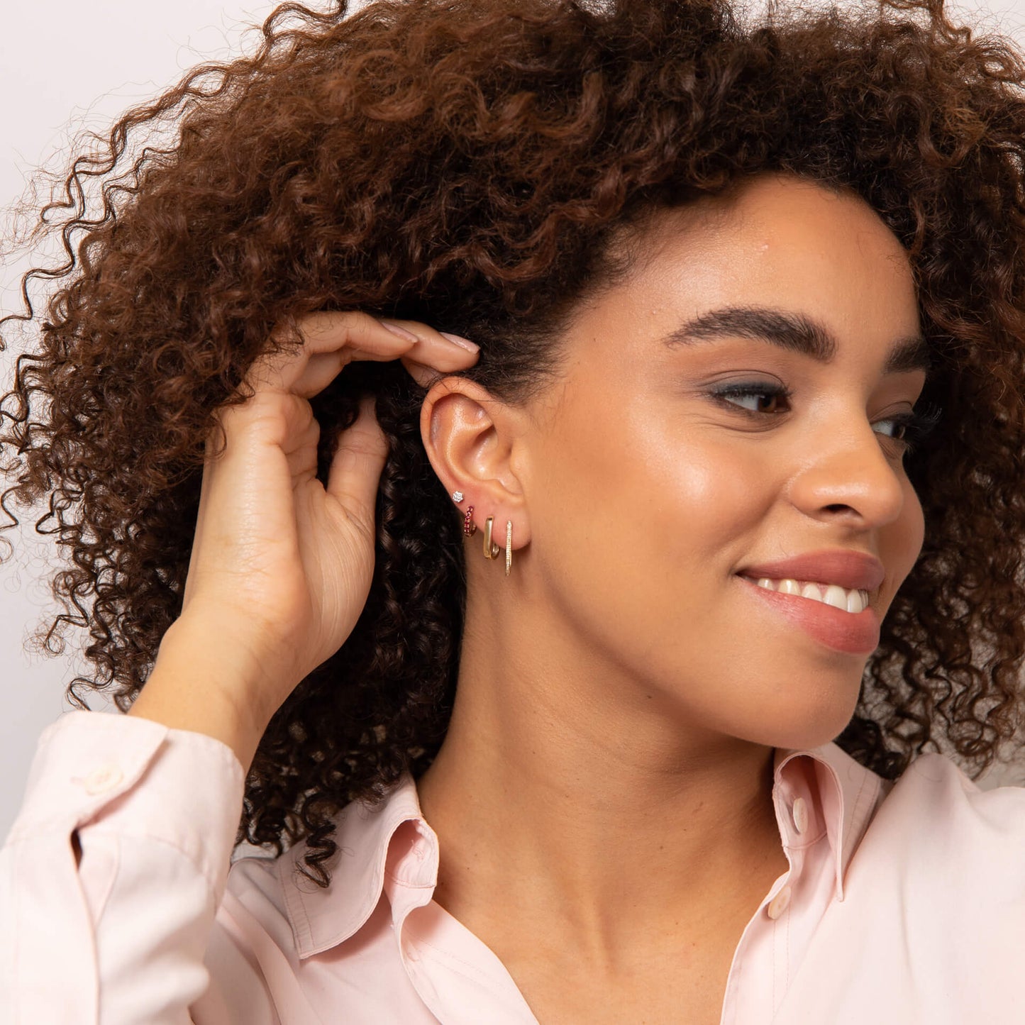 Woman with curly hair wearing gold U hoop earring stacked with other earrings on her ear, wearing a pink shirt against a neutral background