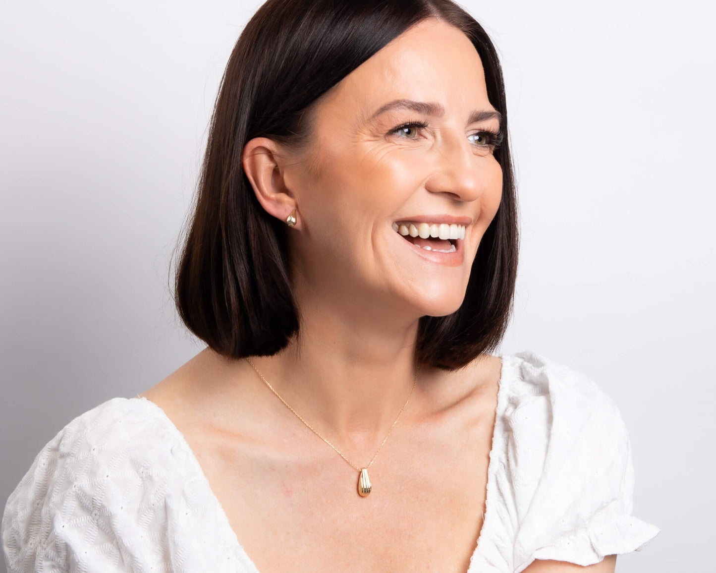 from far Woman wearing a white top and Gold Ridged Teardrop Necklace and earrings against a light gray background