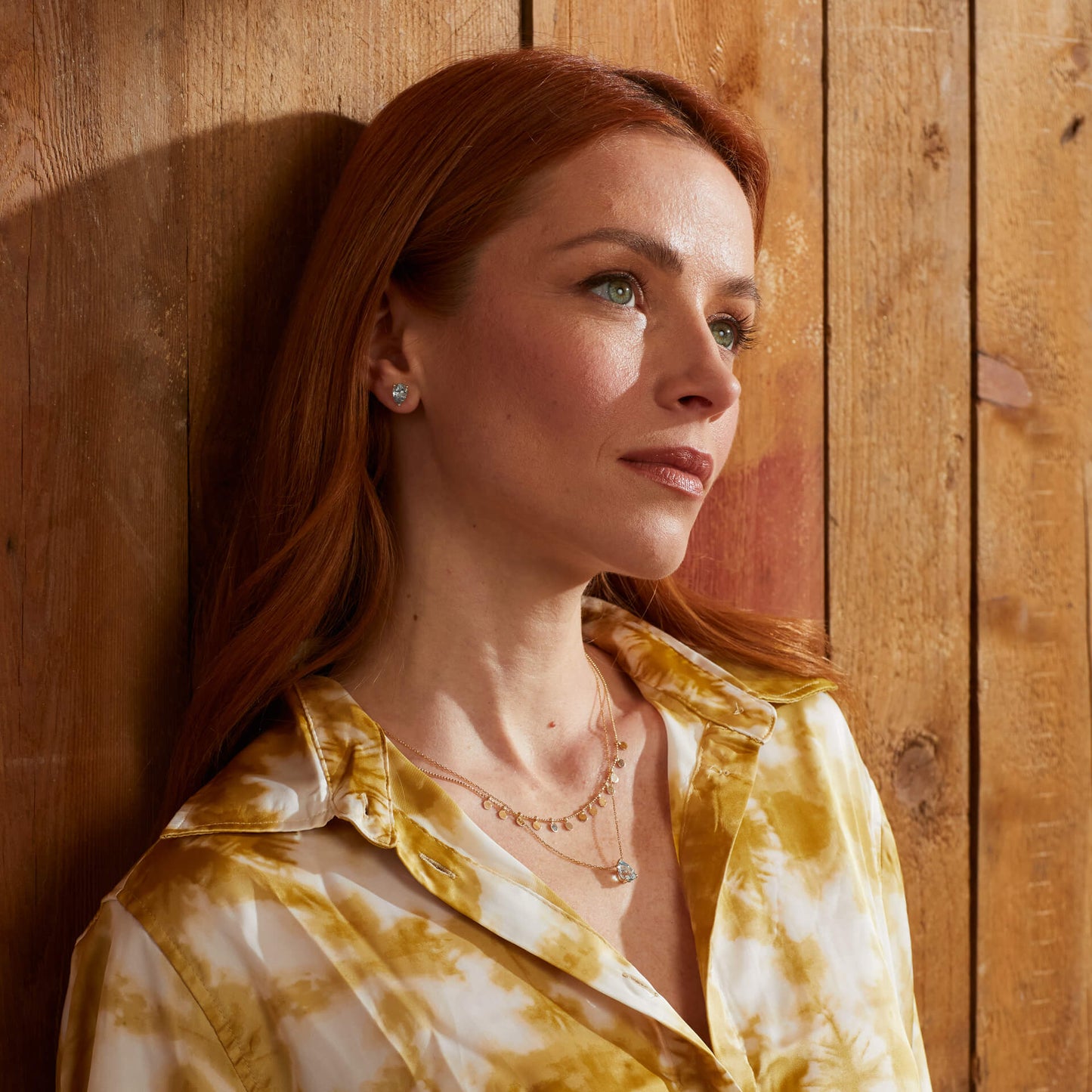 woman wearing Pear Fancy Cut Topaz Gold Stud Earrings and necklace set with another gold necklace layering style. with yellow floral shirt on a wood panel background feature image