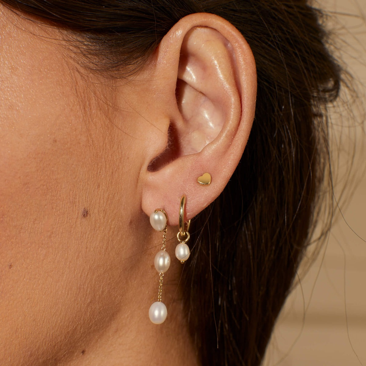 Close-up of an ear wearing gold hoop earrings with pearl drops and a small heart gold earring.