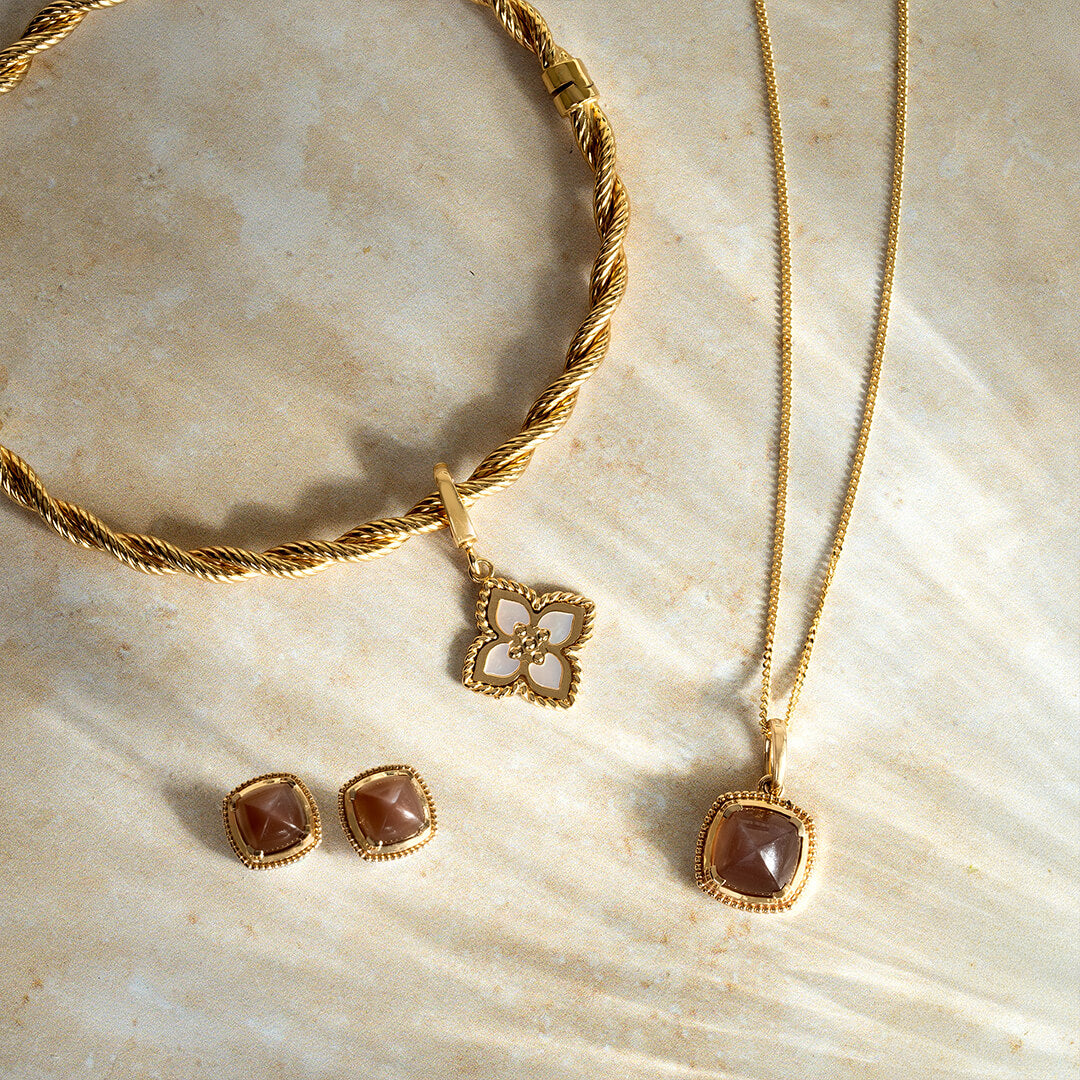 Gold jewellery set featuring a twisted rope bangle, a gold curb chain necklace with a square brown gemstone pendant, and matching square gemstone stud earrings, arranged on a light beige stone surface.