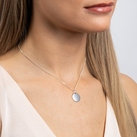 Close-up of a woman wearing a silver engravable pendant necklace with a round pendant on a plain background