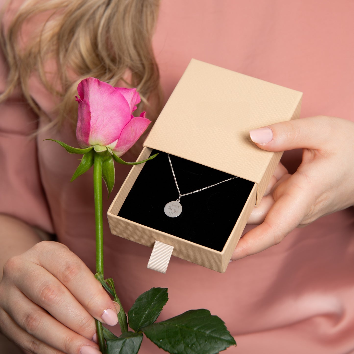 Person holding a pink rose and a jewellery box with a silver engraved necklace inside, on a pink background