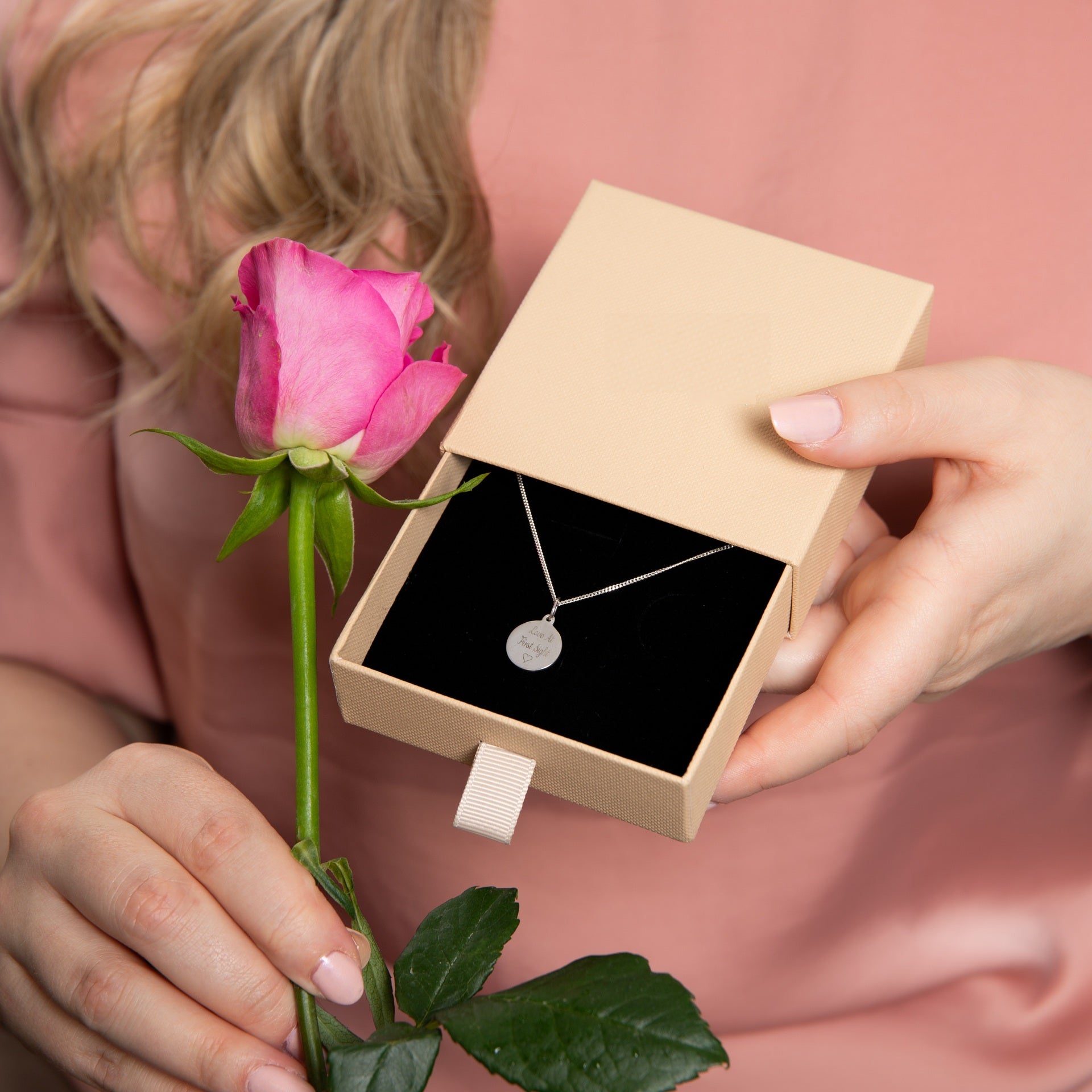 Person holding a pink rose and a jewellery box with a silver engraved necklace inside, on a pink background