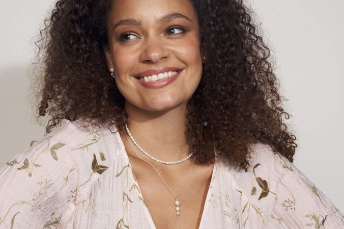 Woman wearing a floral blouse with Rice Pearl Necklace model styling layered necklace style with pearl drop necklace and earrings on a plain background