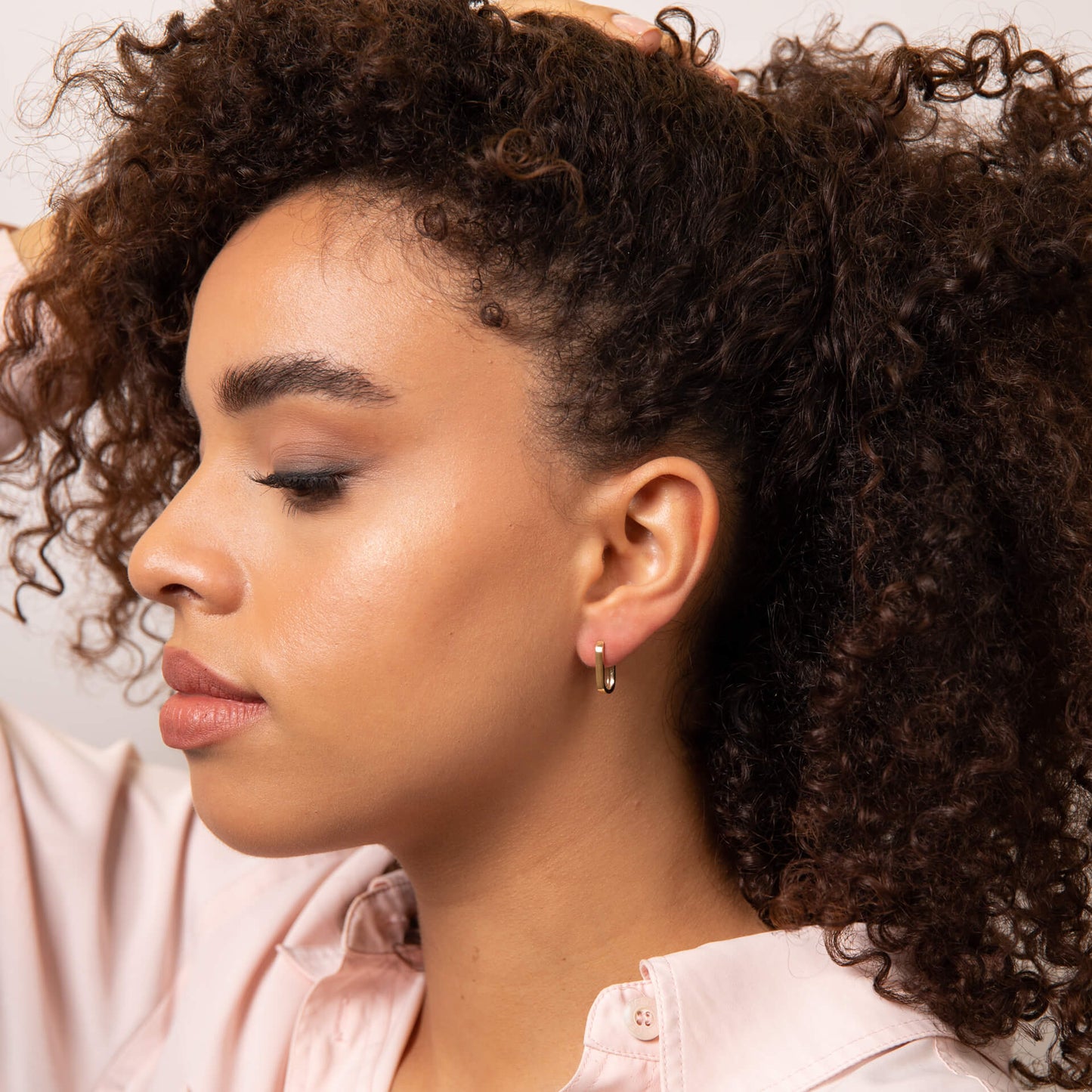 from far ear of a woman with curly hair wearing U hoop gold earring. also wearing a pink shirt.