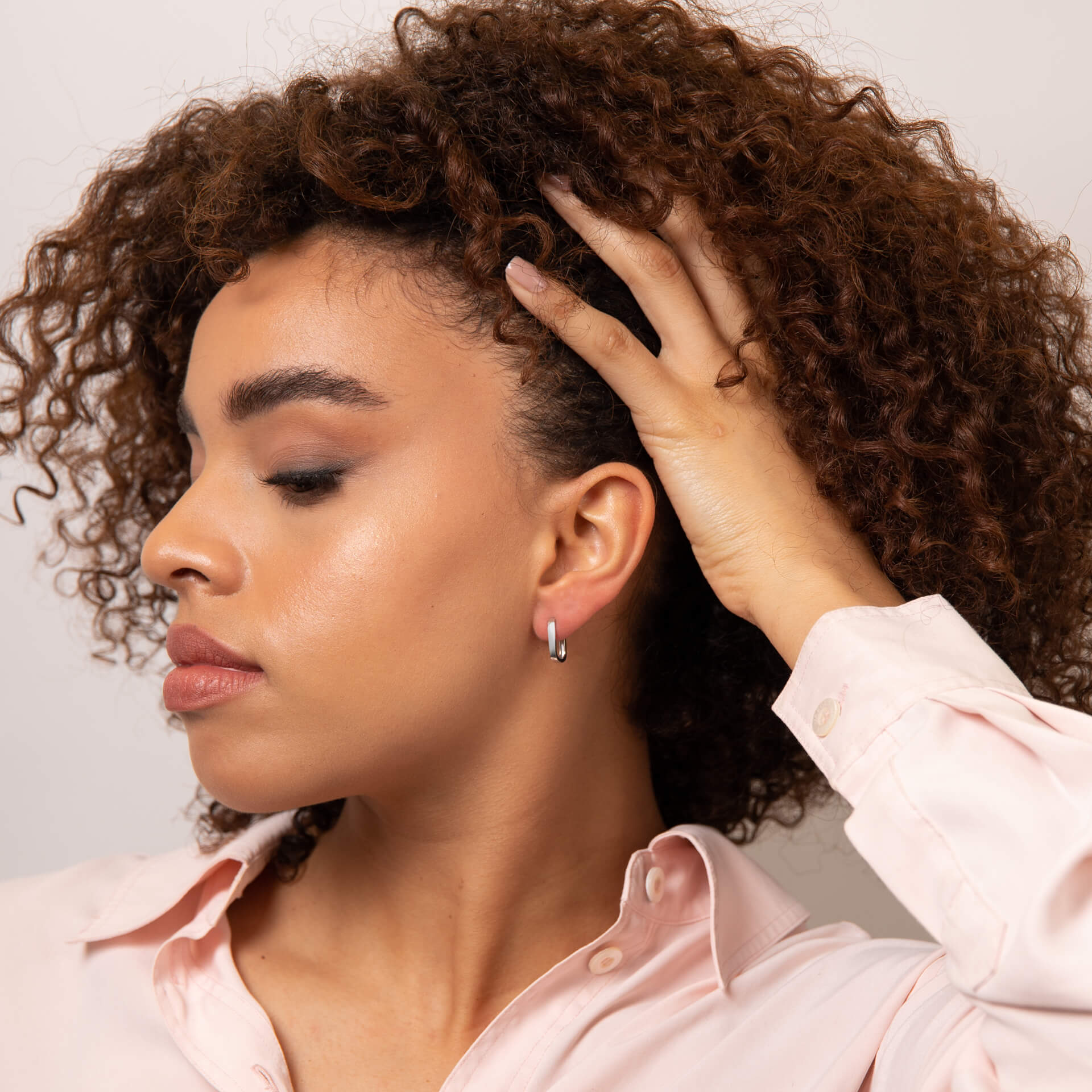 from far Woman with curly hair showing ear wearing a white gold U hoop earring with a pink shirt against a neutral background.