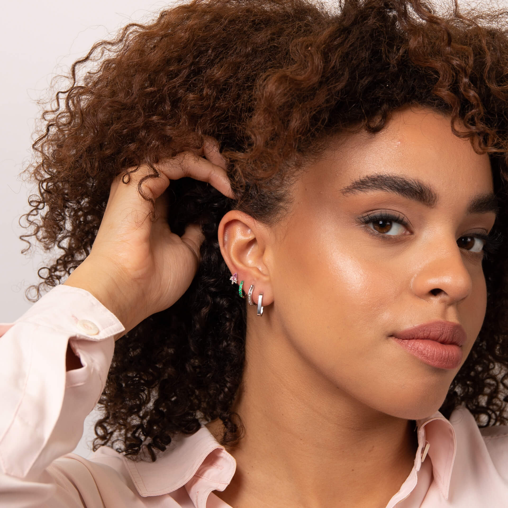 from far Woman with curly hair showing her ear wearing white gold U hoop earring stacked with other earrings and wearing a pink shirt against a light background