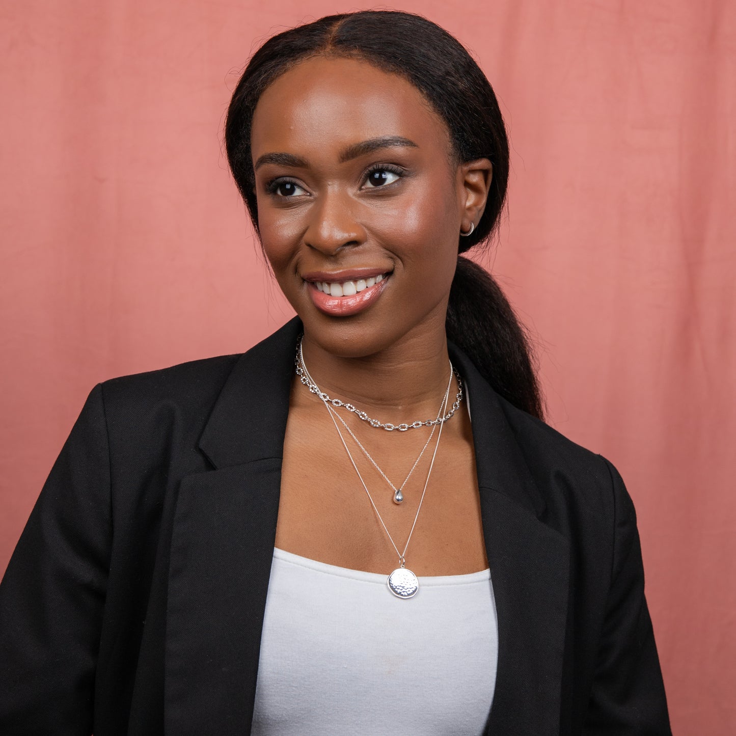 Woman wearing a black blazer and white top with style example layered Solid Teardrop Necklace silver. layered silver necklaces arounf her neck and shes standing against a pink background
